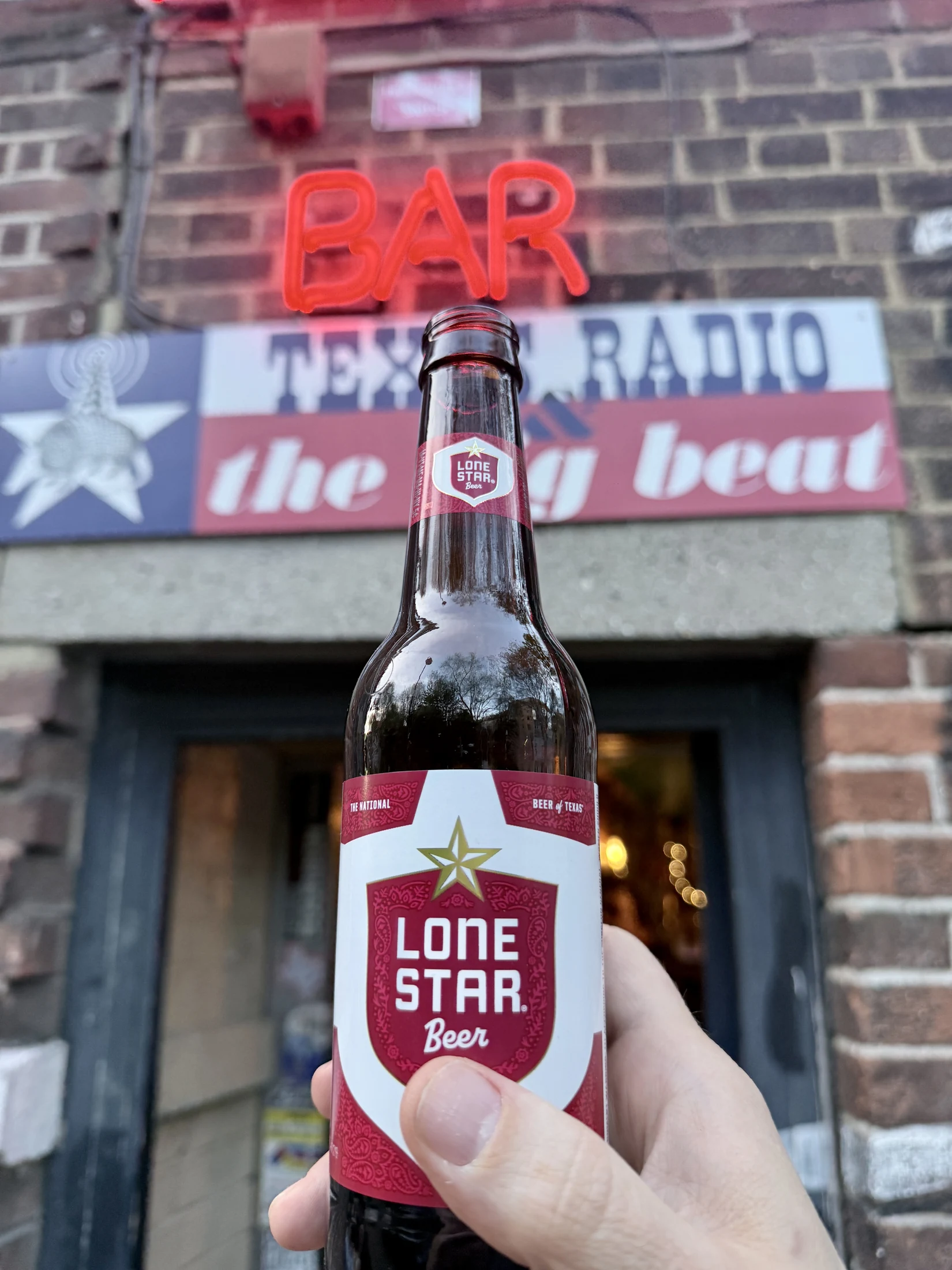 A bottle of Lone Star Beer outside the Flying W, beneath the red BAR neon and Texas Radio sign