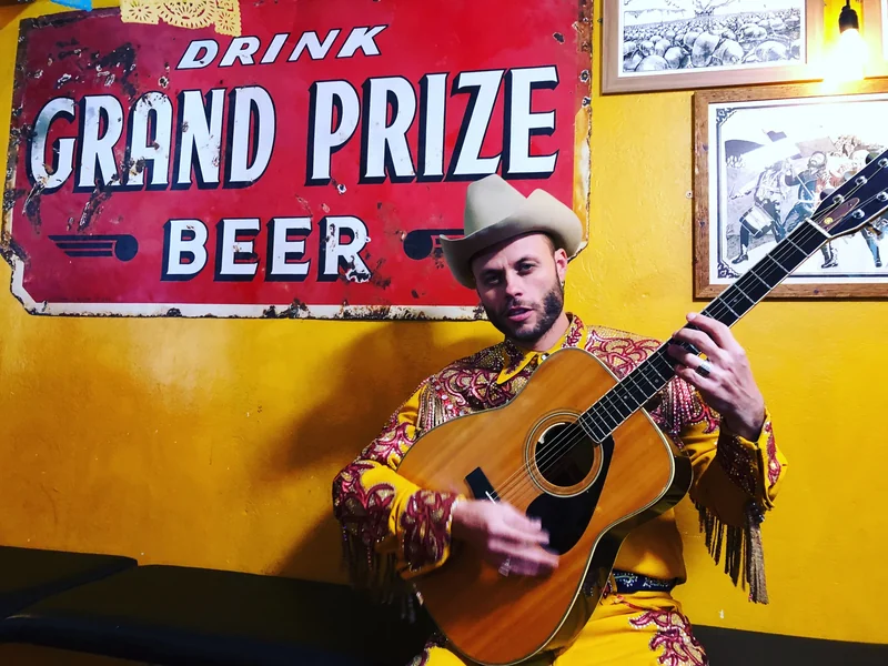 Charley Crockett with his guitar beneath a vintage Grand Prize beer poster